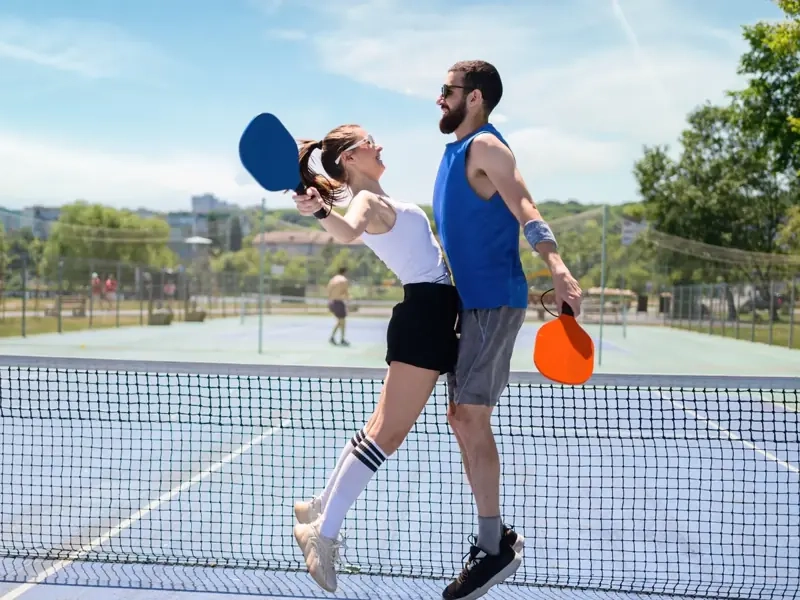A happy couple jumping together at the net, celebrating a point on a sunny pickleball court