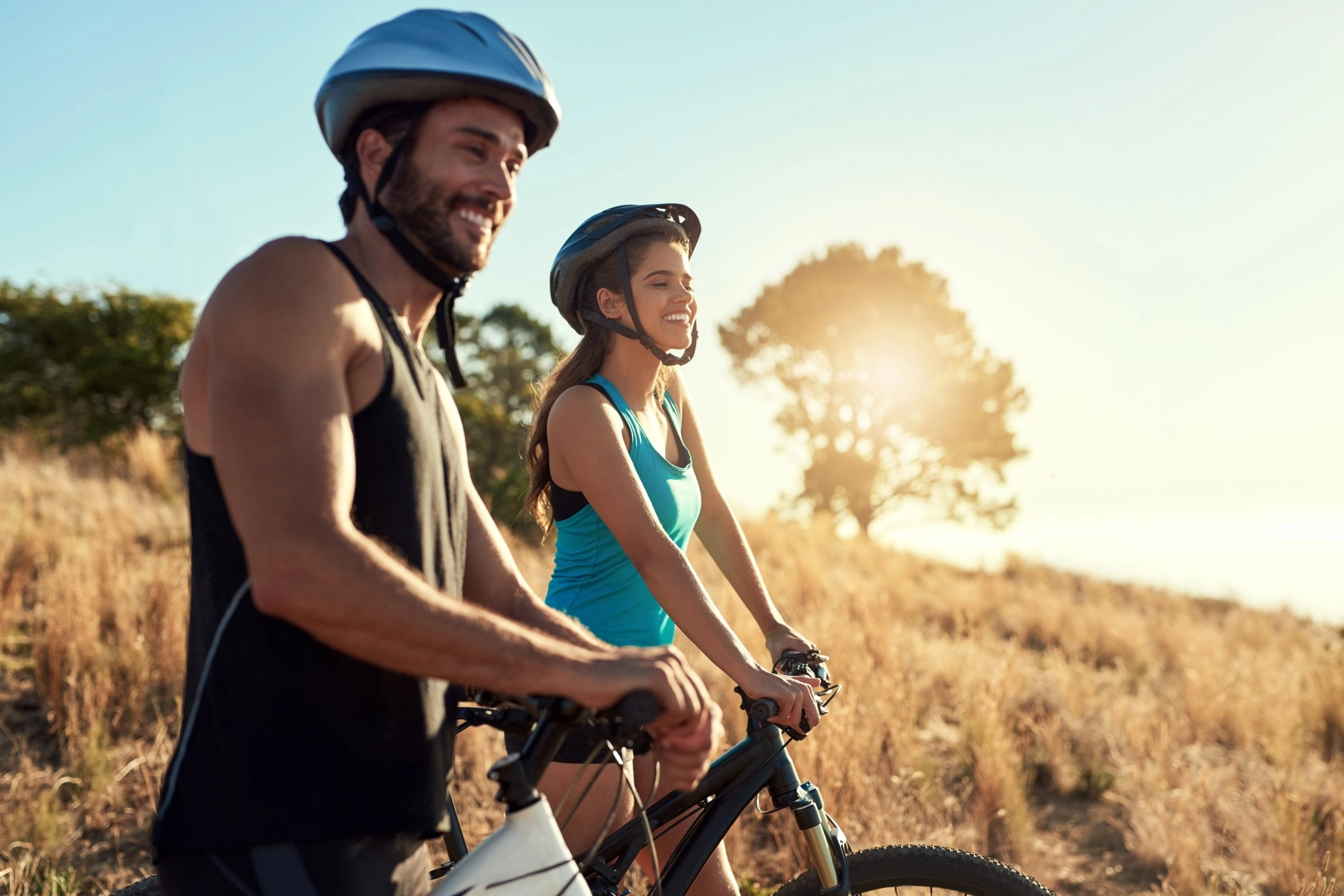 A smiling man and woman in cycling helmets with their mountain bikes on a sunny grassy hillside