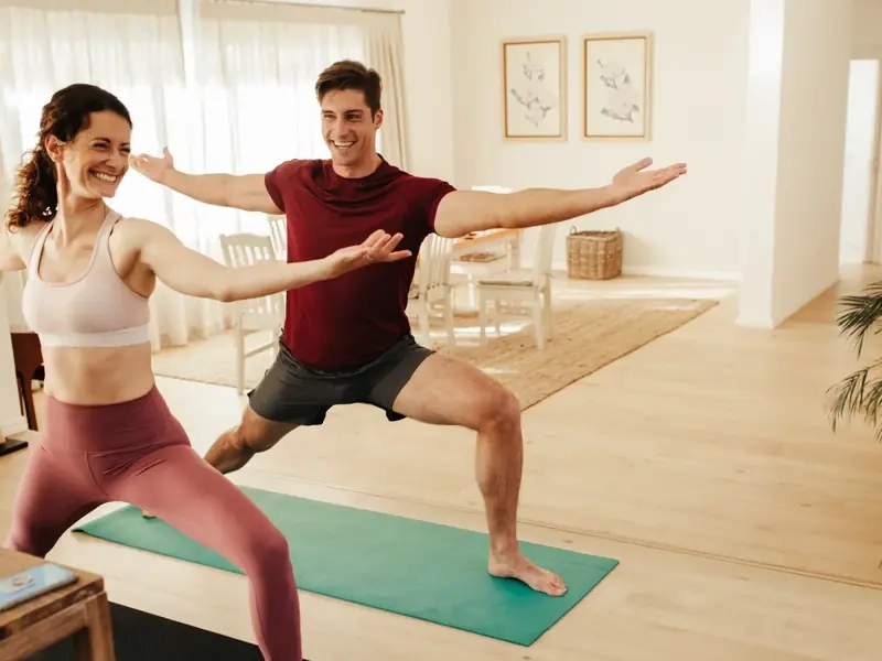 A smiling couple holding warrior pose side by side on yoga mats in a bright living room