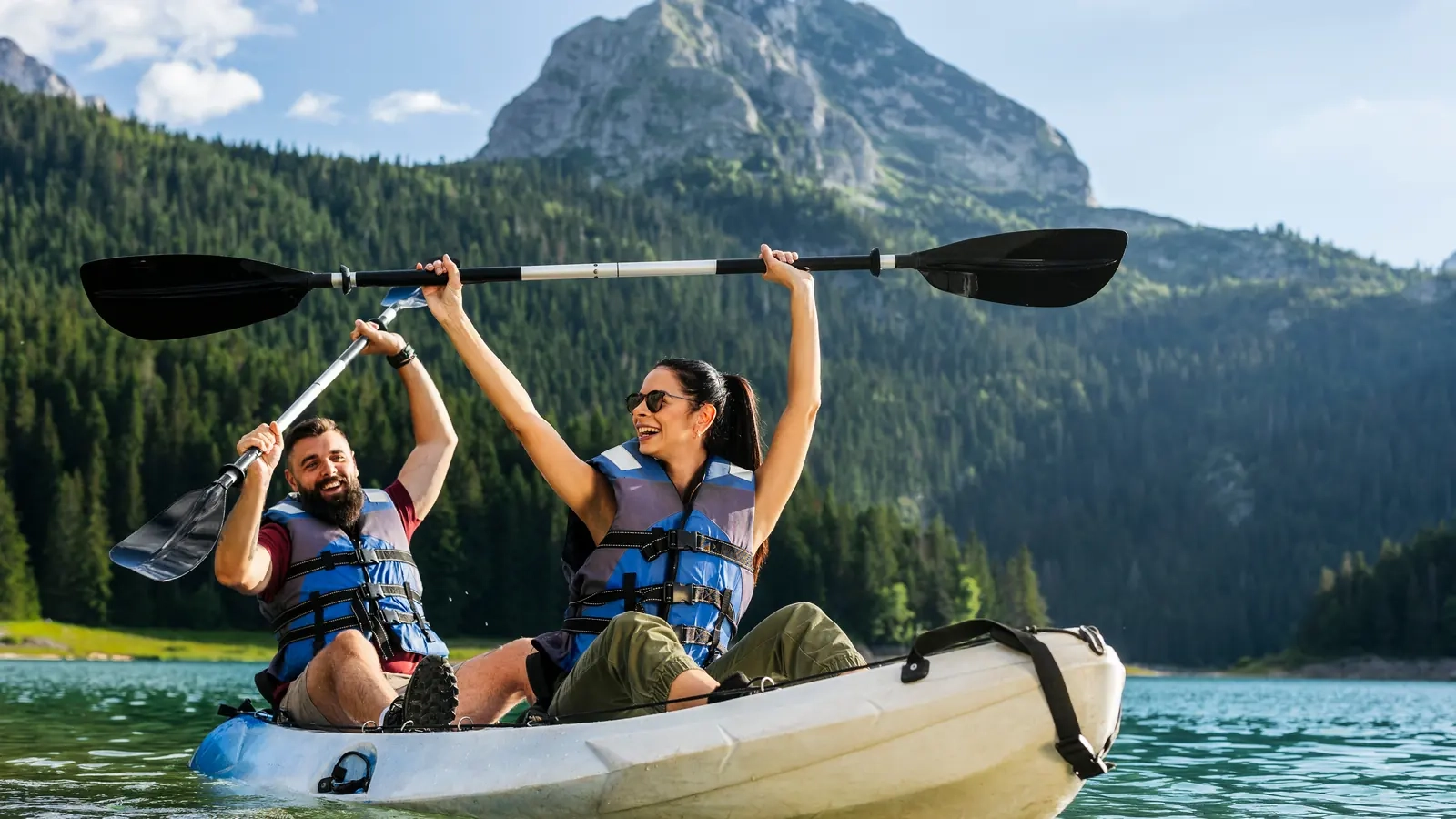 A couple in lifejackets sitting in a tandem kayak on a mountain lake, both raising their paddles and smiling