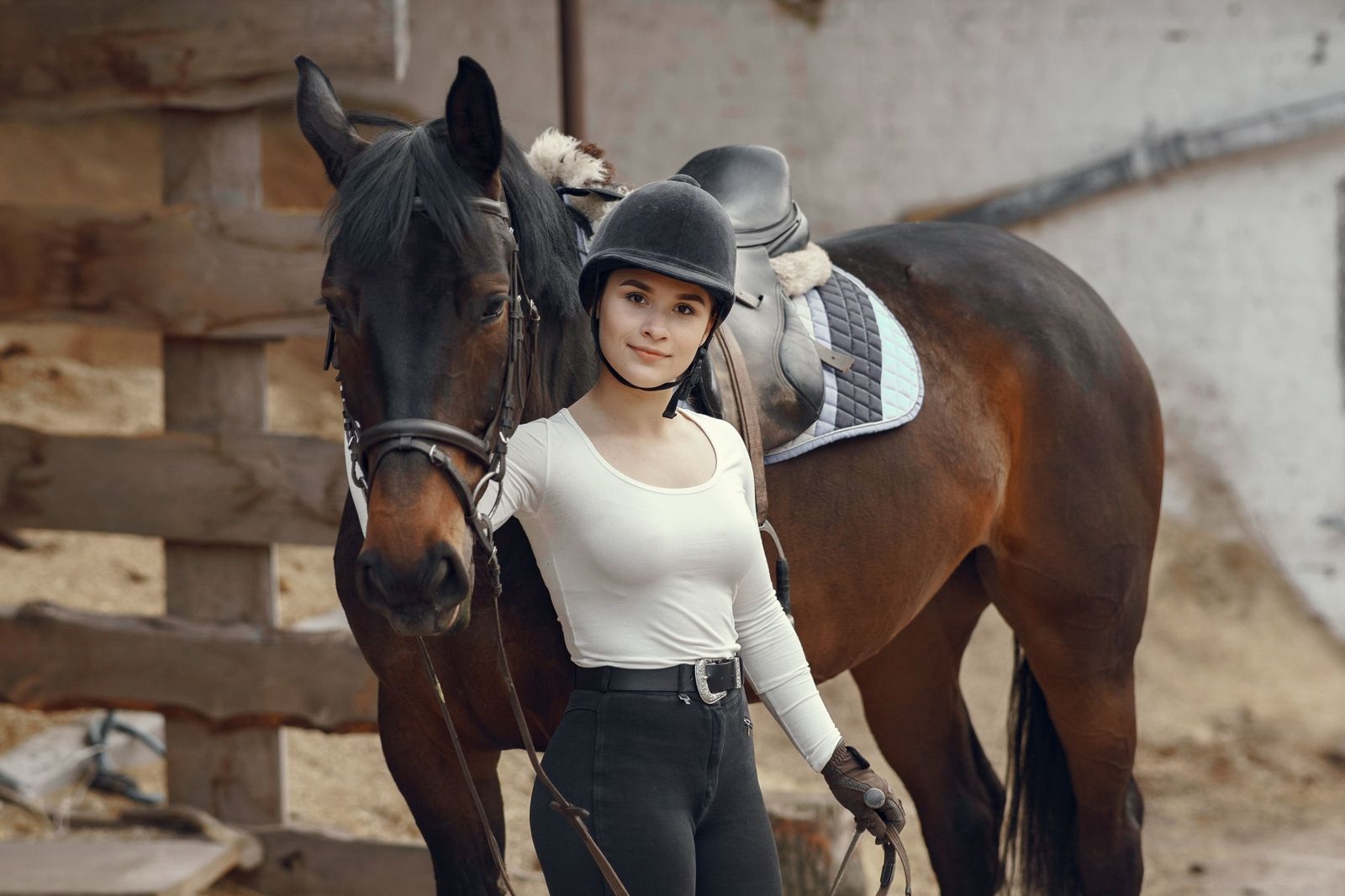 A young rider in a velvet riding hat standing beside her saddled bay horse at the stable