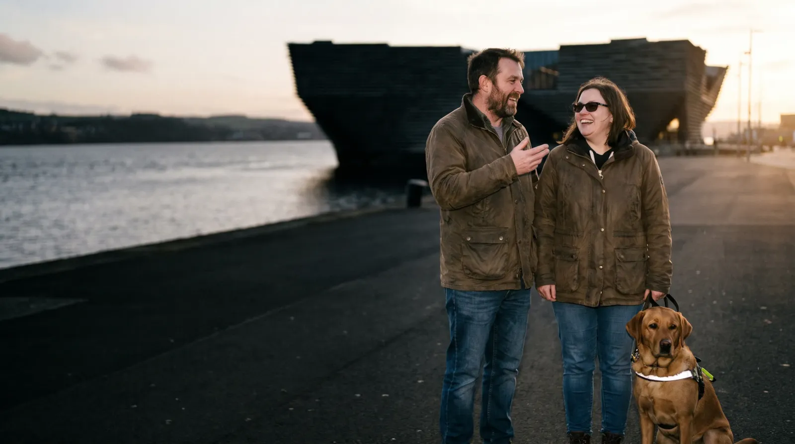 A couple laughing on the Dundee waterfront beside the V&A at dusk; the woman is visually impaired, with her guide dog