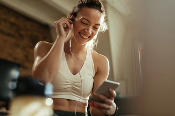 A smiling woman in workout clothes adjusting her earphones while looking at her phone