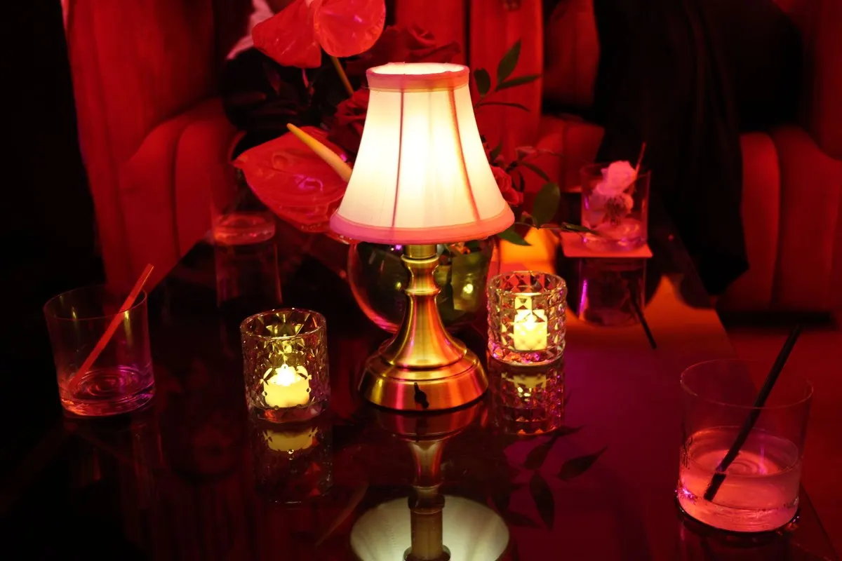 A glowing table lamp, candles and red flowers in a cozy lounge with deep red curtains