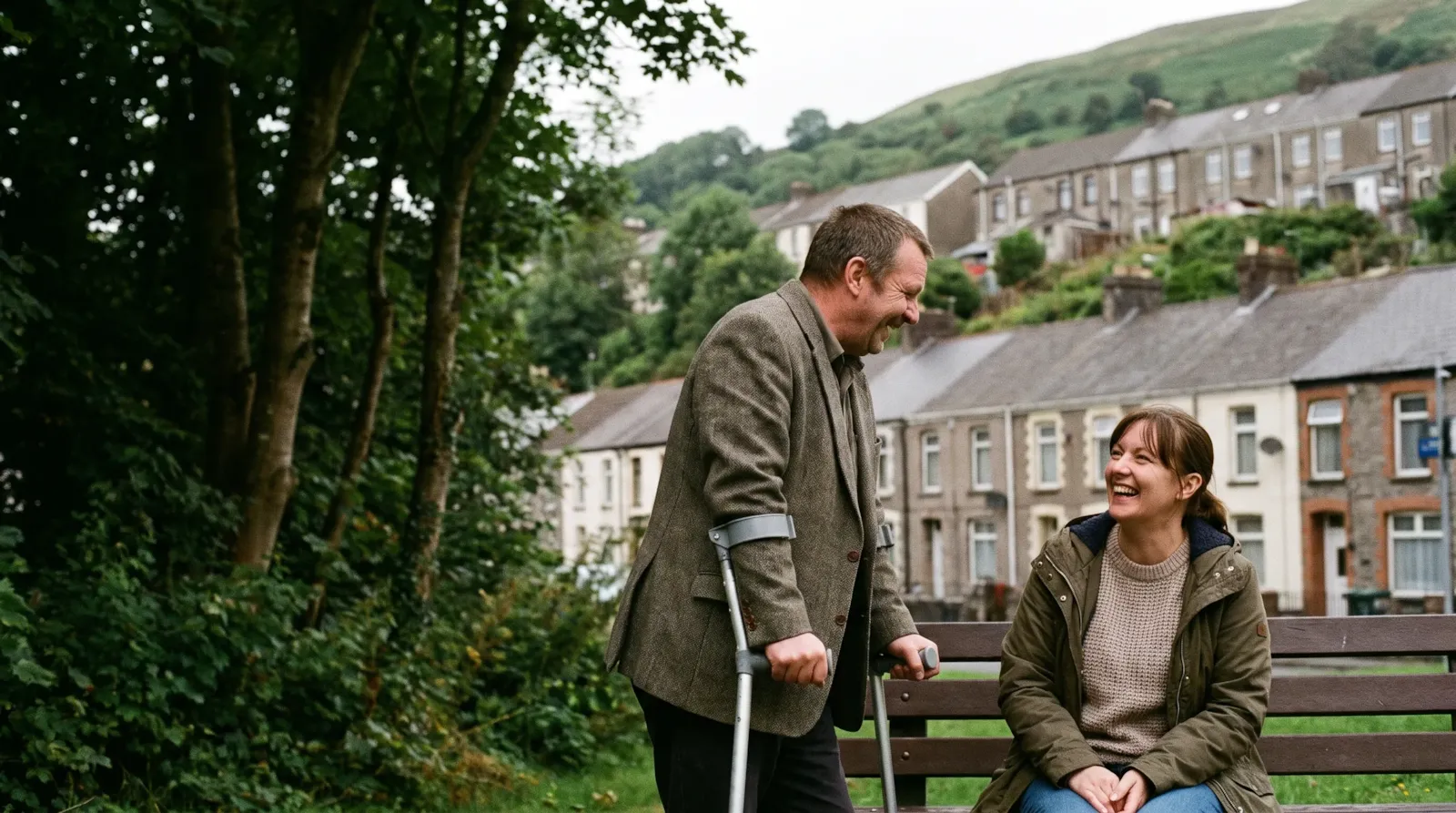 A couple laughing in a valley park with terraced houses on the hillside; the man uses forearm crutches