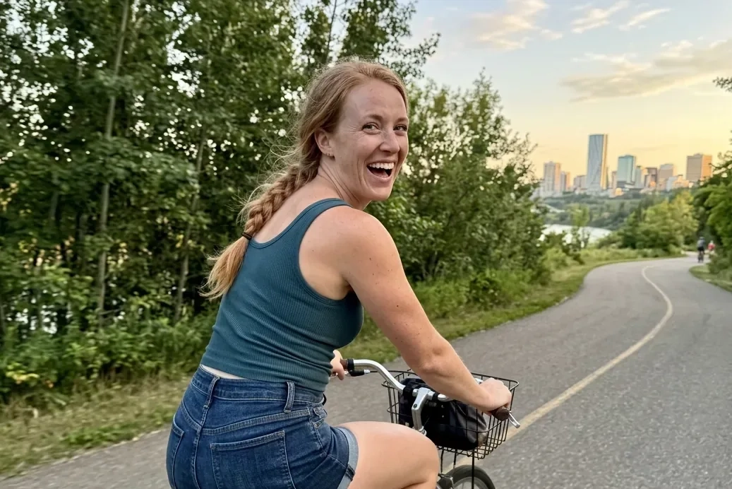 A laughing woman with a long strawberry-blonde braid, a fitted tank top and denim shorts rides a bicycle along a leafy river valley trail and looks back over her shoulder, with the Edmonton skyline behind her (AI-generated image)