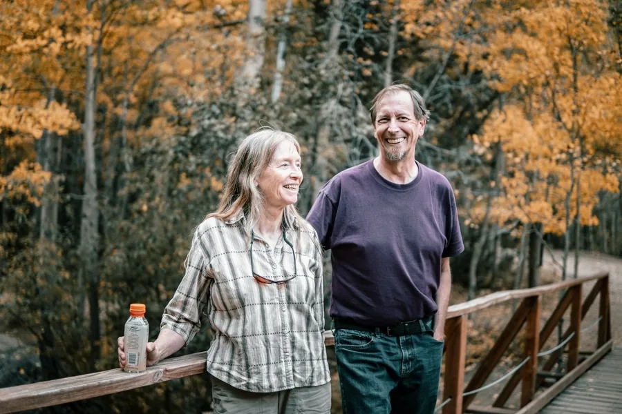 A smiling man and woman standing by a wooden footbridge among golden autumn trees