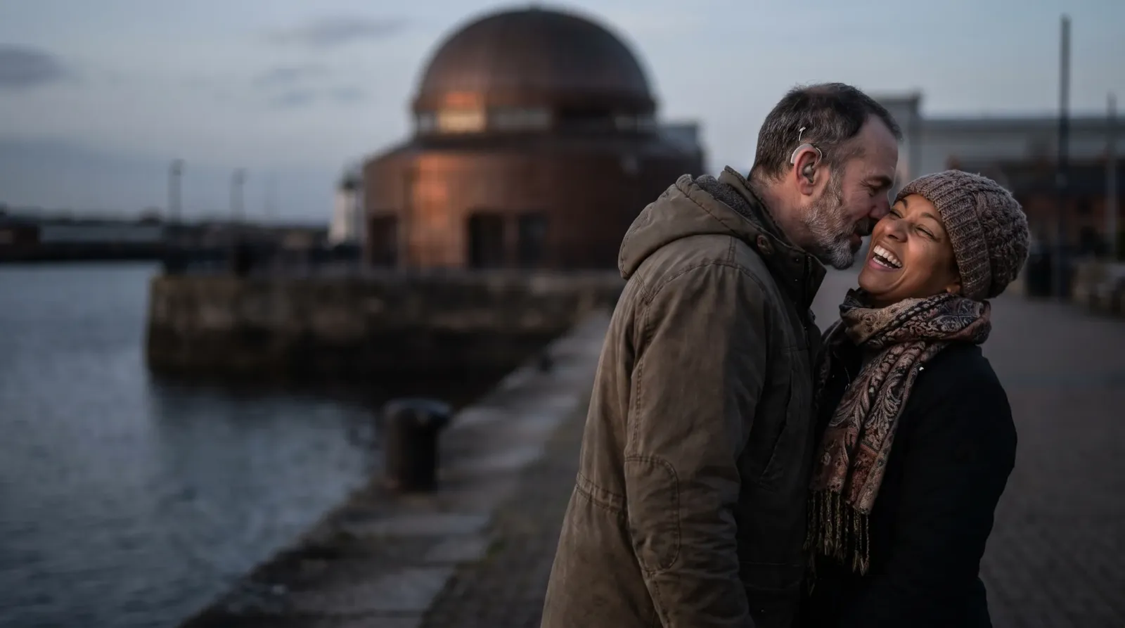 A couple laughing on the Cardiff Bay waterfront at dusk; the man wears hearing aids
