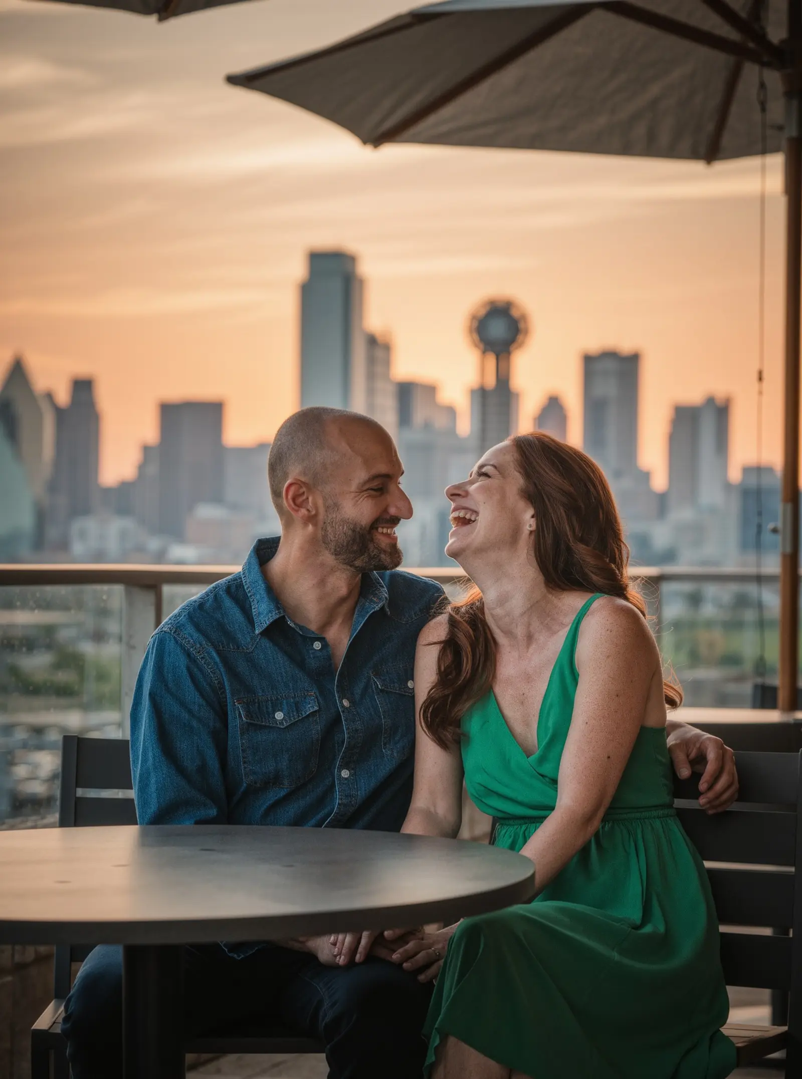 A couple laughing together on a rooftop patio at sunset with the Dallas skyline behind them
