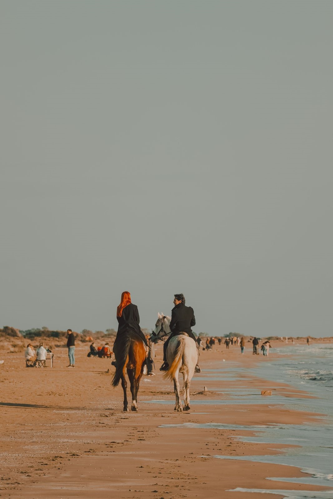 Two riders walking their horses along a wide sandy beach at low tide under a grey sky