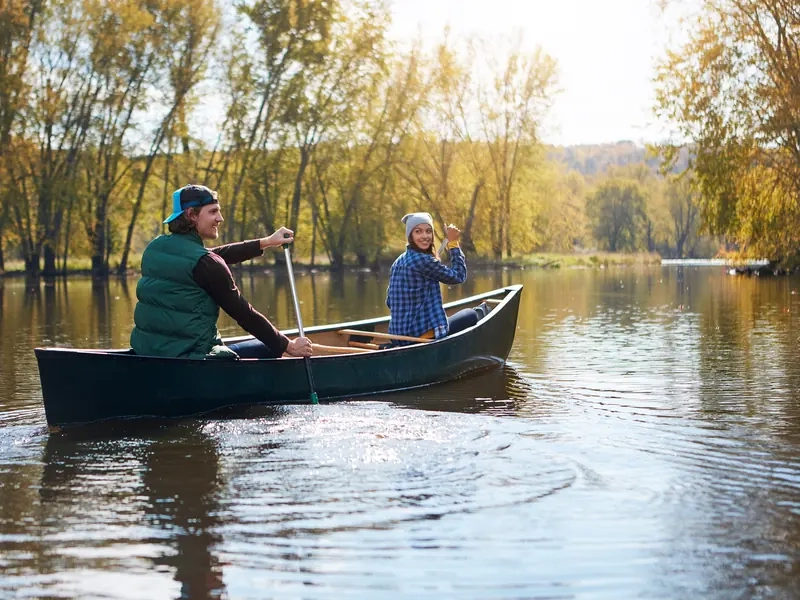 A couple paddling a canoe together across a calm lake lined with autumn trees