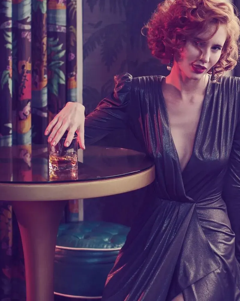 A red-haired woman in a plunging metallic dress leaning on a bar table with a drink, giving the camera a sultry look