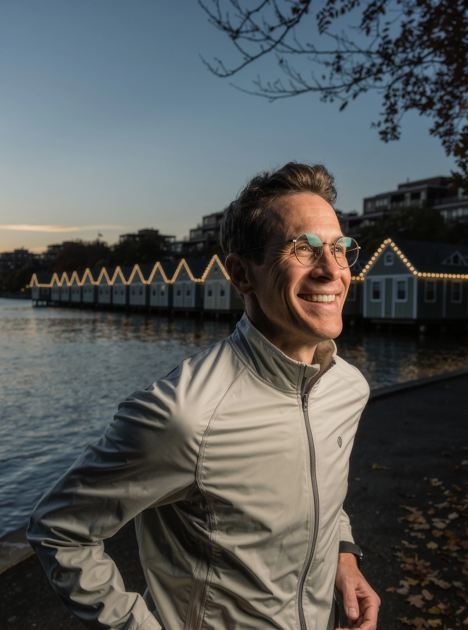 A smiling man in glasses and a running jacket by the river at dusk, with a row of lit boathouses behind him