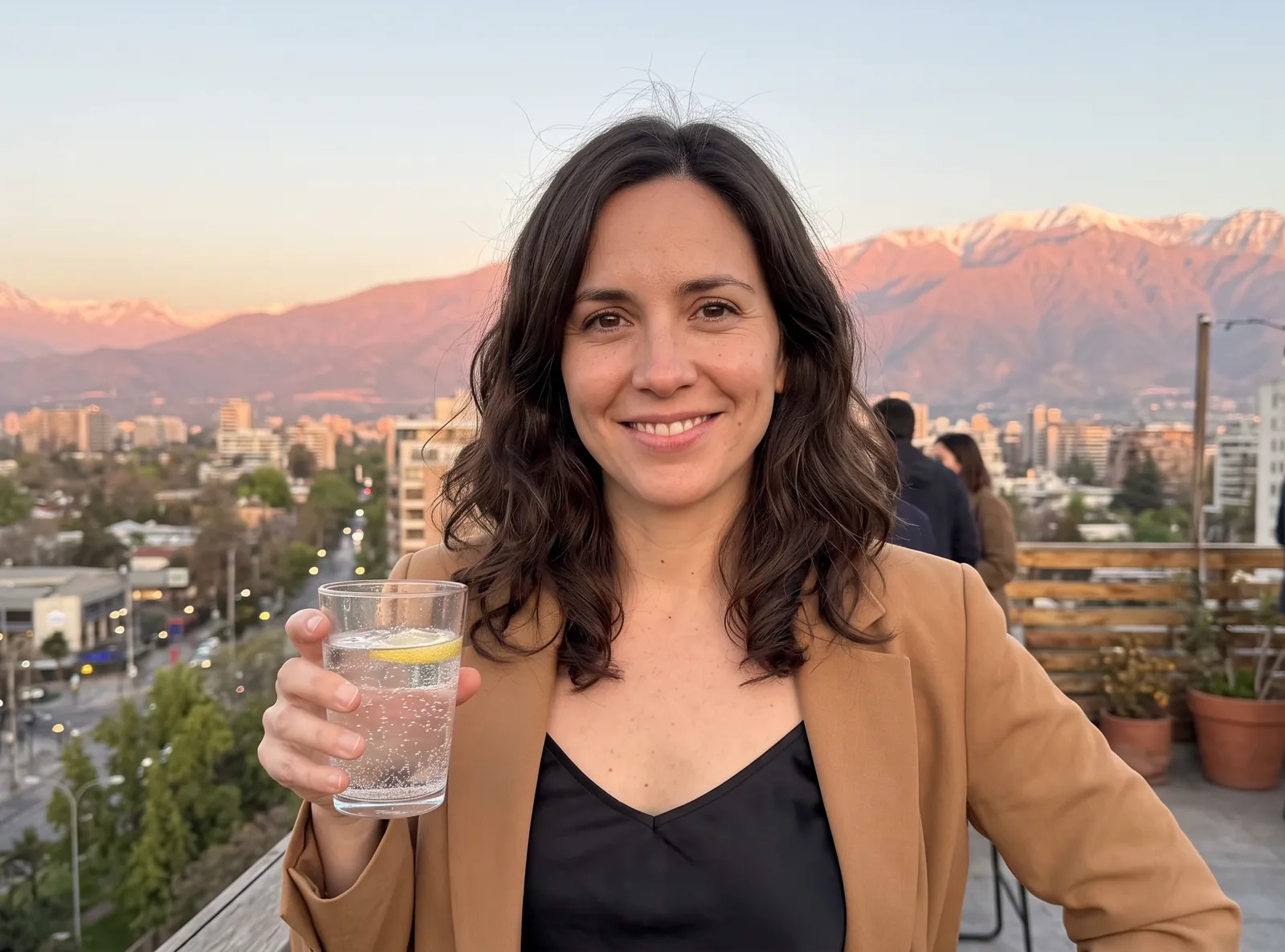 Una mujer con pelo castaño ondulado, camisola negra y blazer camel sonríe en una terraza de Santiago al atardecer, con la cordillera nevada de los Andes detrás (imagen generada por IA)