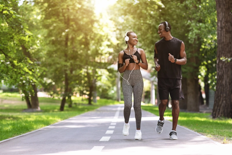 A smiling woman and man in headphones jogging side by side along a leafy park path