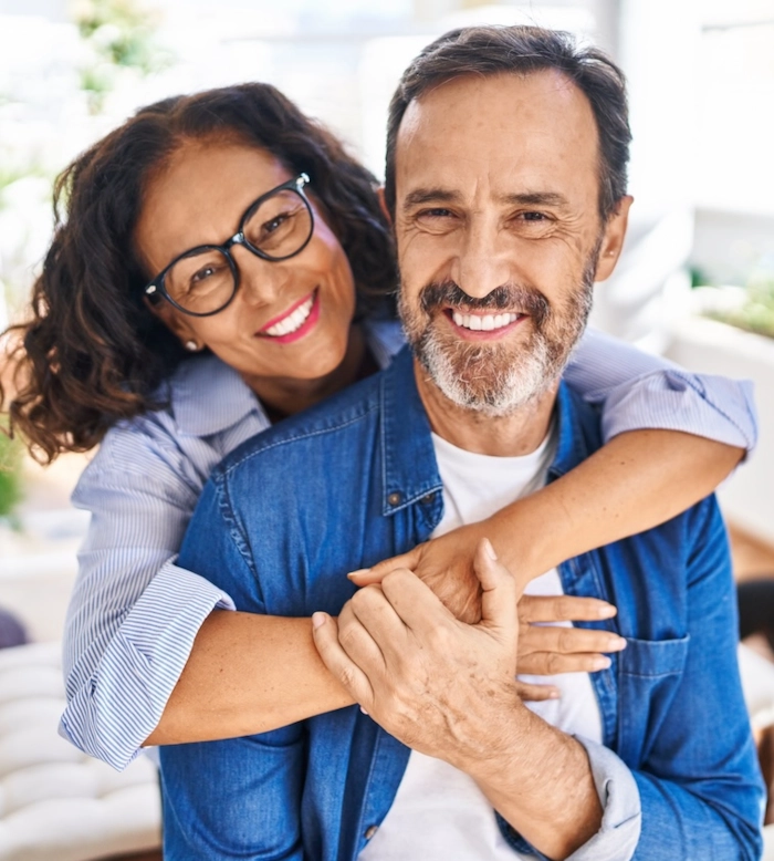 A couple in their fifties, she hugging him from behind, both smiling