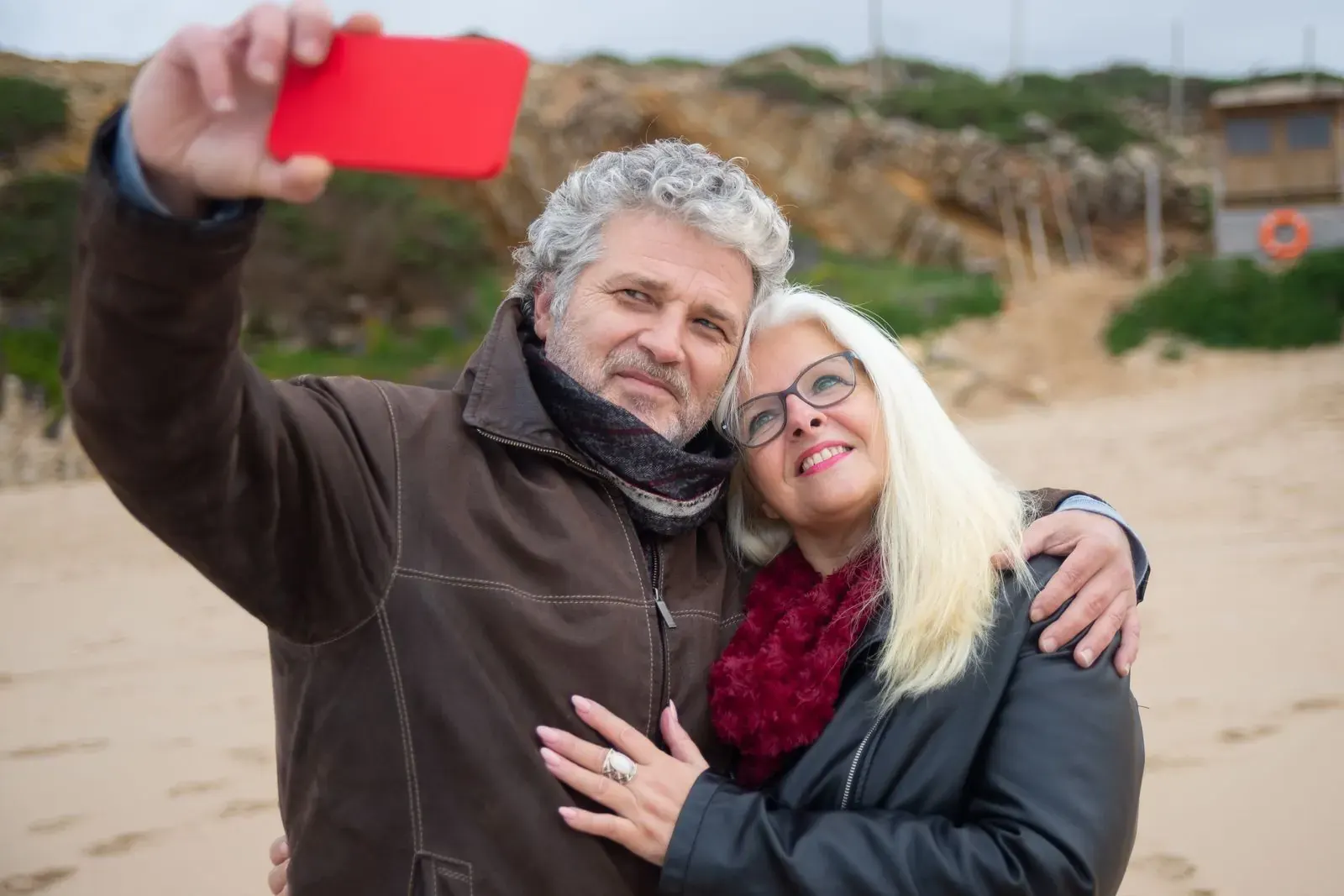 A laughing couple taking a selfie with a red phone on a sandy beach path