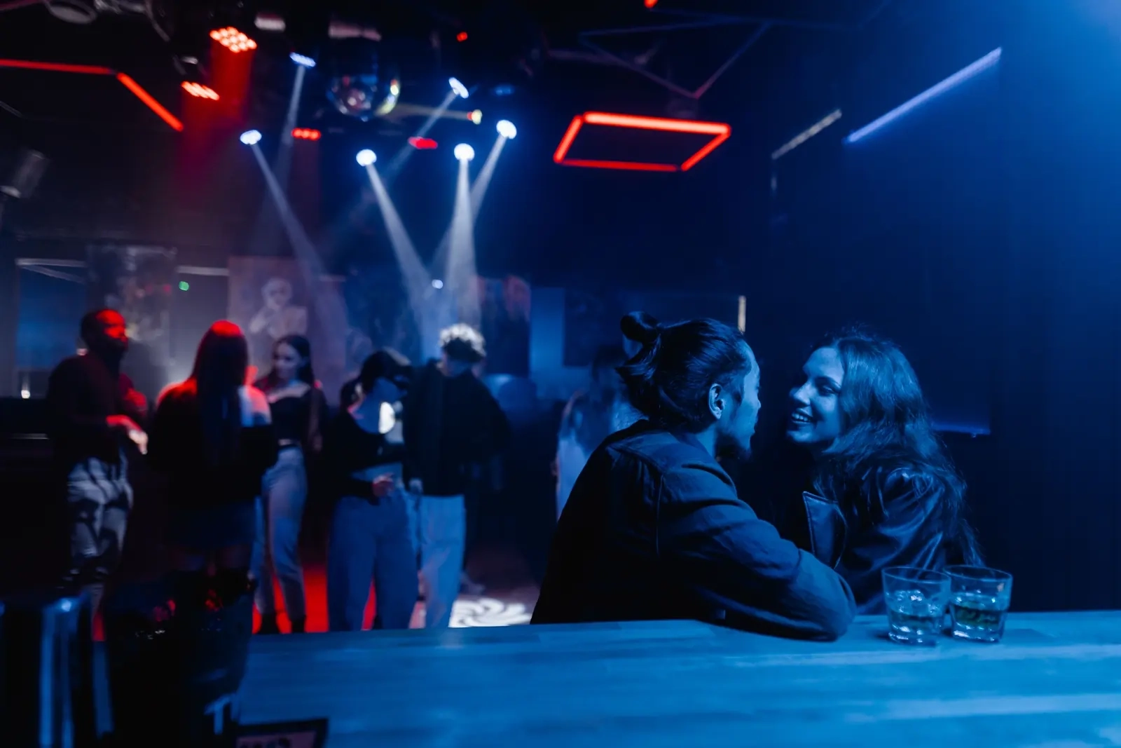 A crowded nightclub dance floor under blue stage lights, with a couple talking at the bar, illustrative photo of models