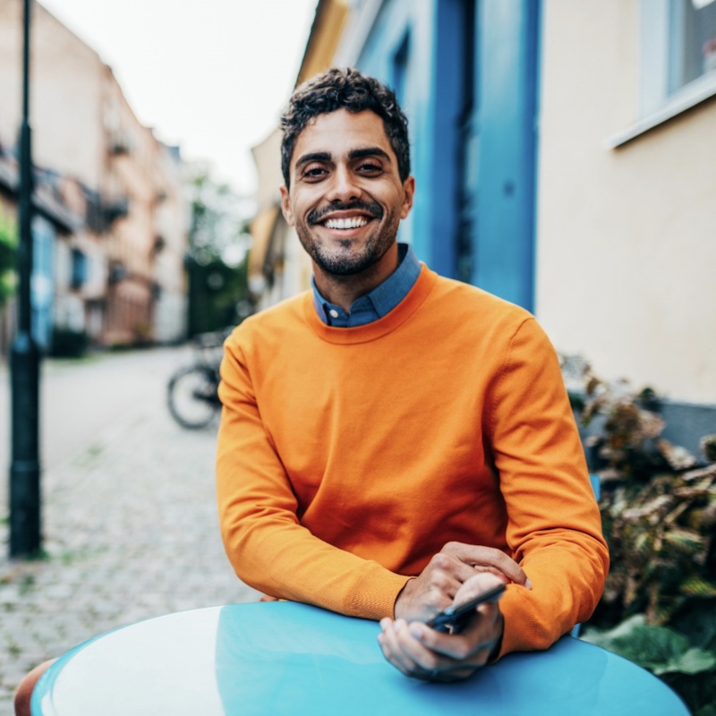 A man in an orange sweater smiles with his phone in hand, on a terrace
