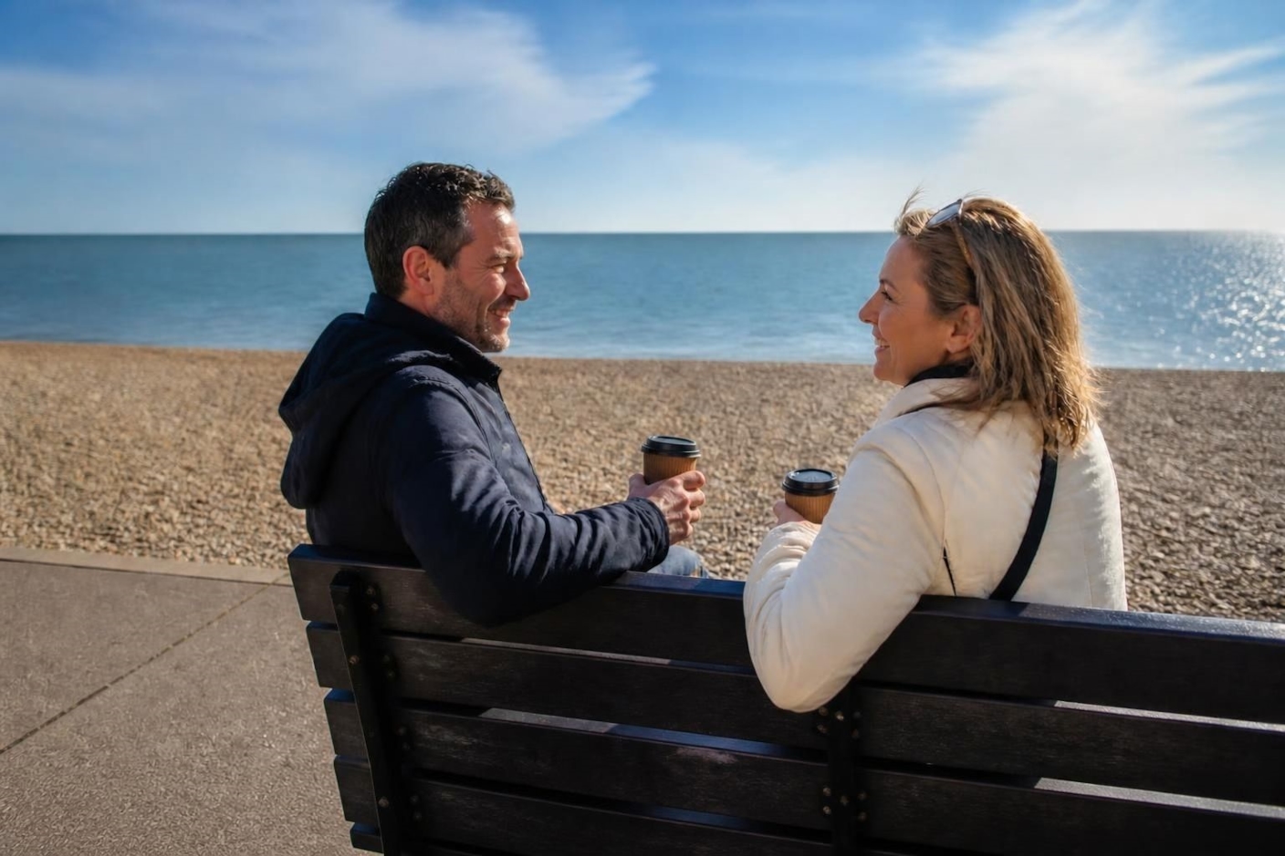 A couple laughing over takeaway coffees on a seafront bench, with a pebble beach and calm sea behind them