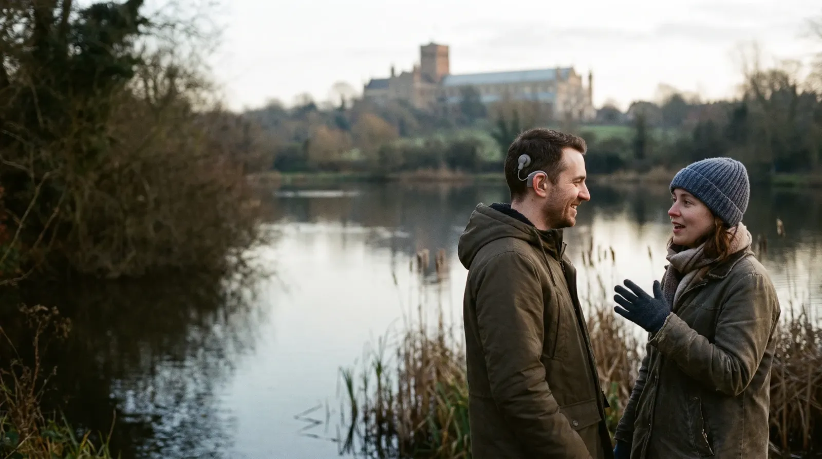 A couple chatting by the lake in Verulamium Park, St Albans, with the cathedral behind; the man has a cochlear implant