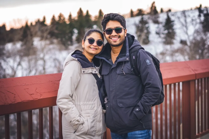 Smiling couple in sunglasses and winter jackets on a boardwalk with snowy pines behind them
