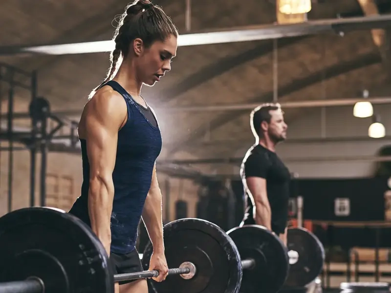 A woman and man setting up for barbell deadlifts in a warm, sunlit gym