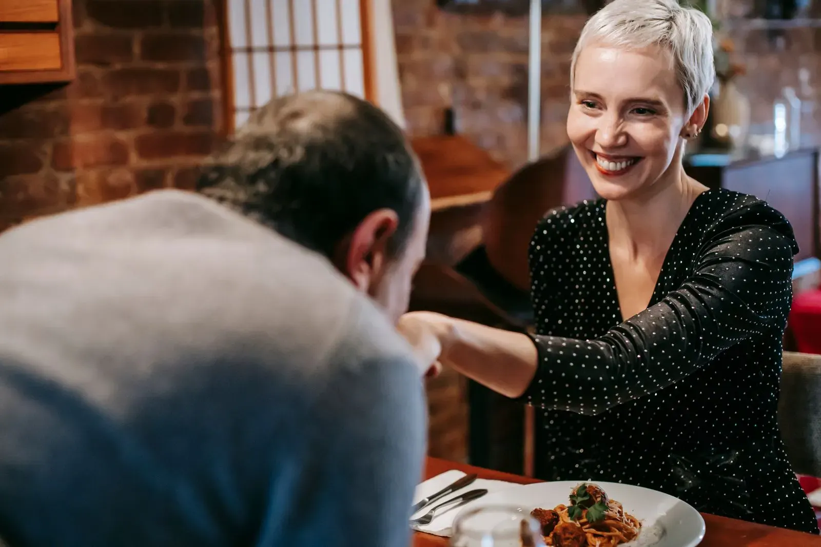 A smiling woman with short grey hair at a restaurant table while her date kisses her hand