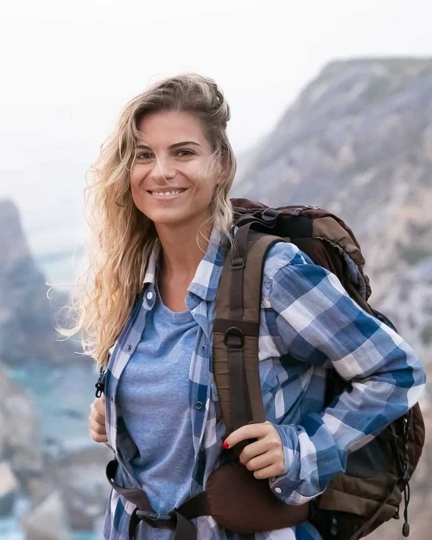A smiling woman in a plaid shirt with a backpack on a coastal hiking trail, cliffs behind her
