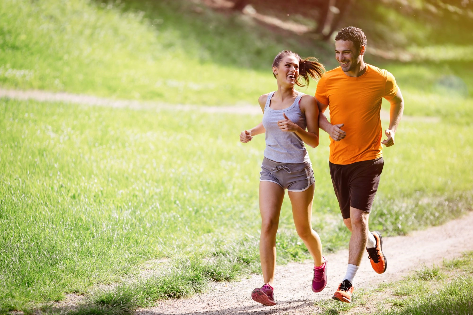 A laughing woman and man jogging side by side along a sunny meadow path