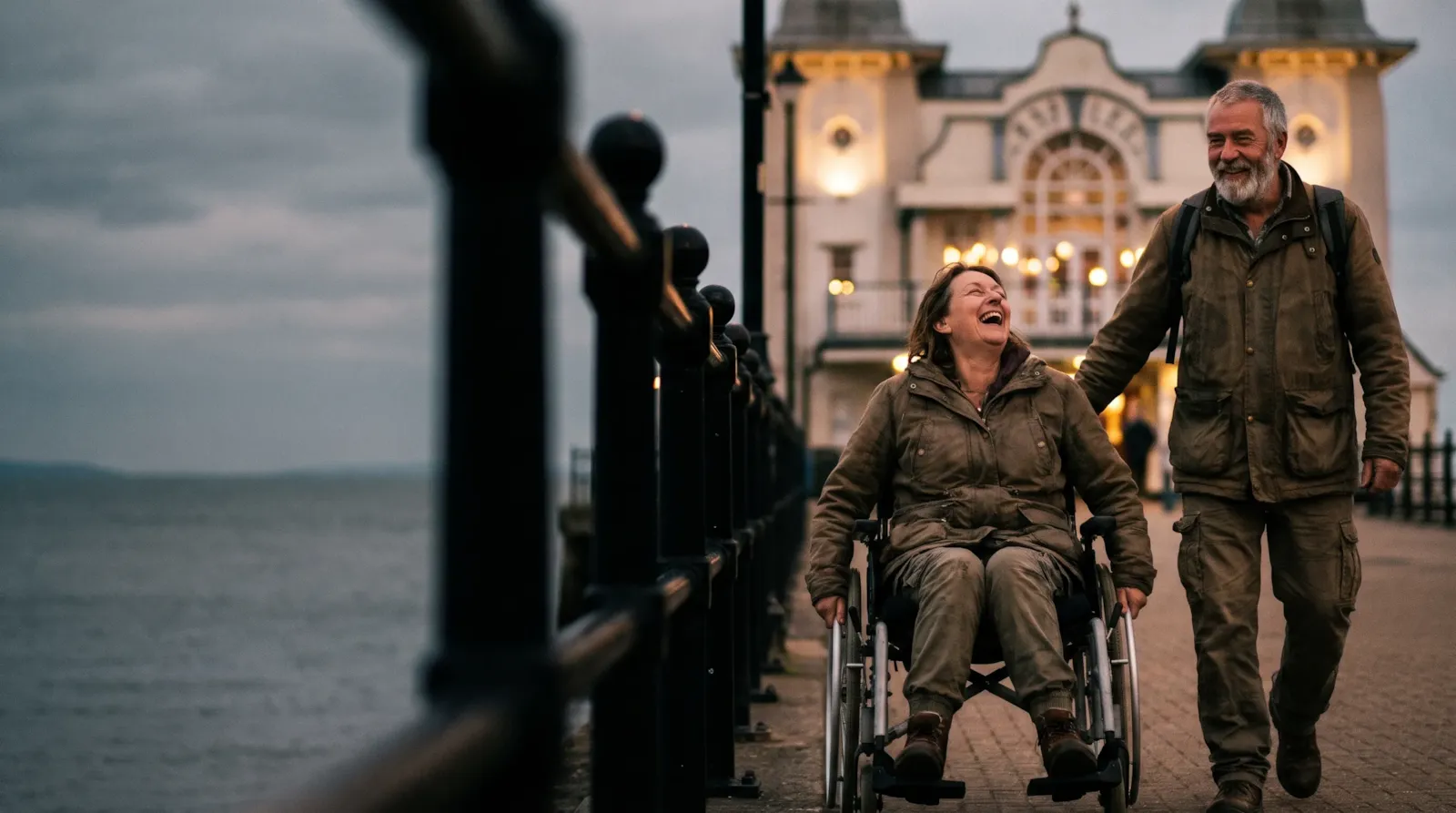 A couple laughing on the seafront at Penarth by the lit pier pavilion; the woman is a wheelchair user