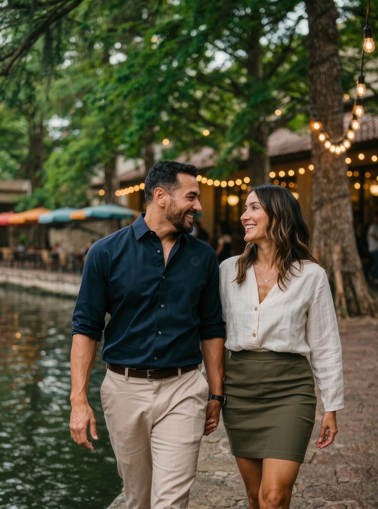 A smiling couple in their forties walking beside a river lined with trees and string lights