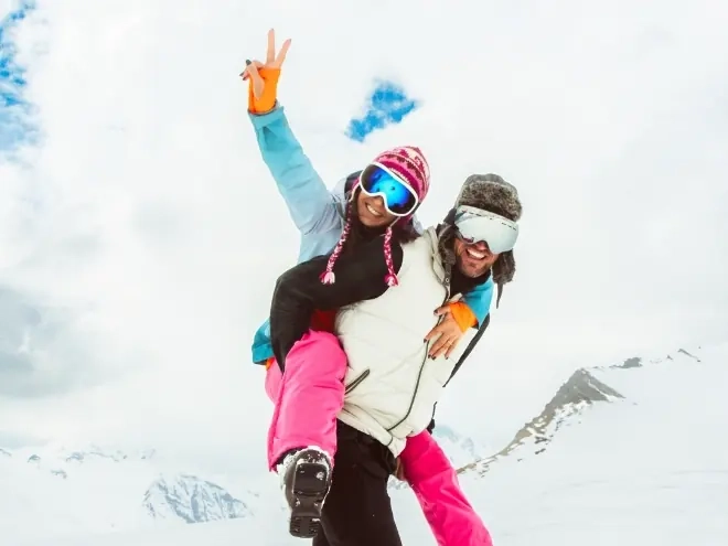 A woman sitting on a man's shoulders in the snow, both raising their arms and laughing