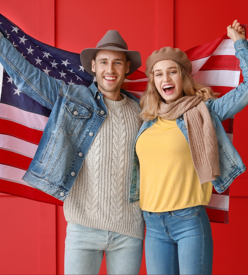 A happy couple in denim jackets and hats holding the American flag up against a red wall