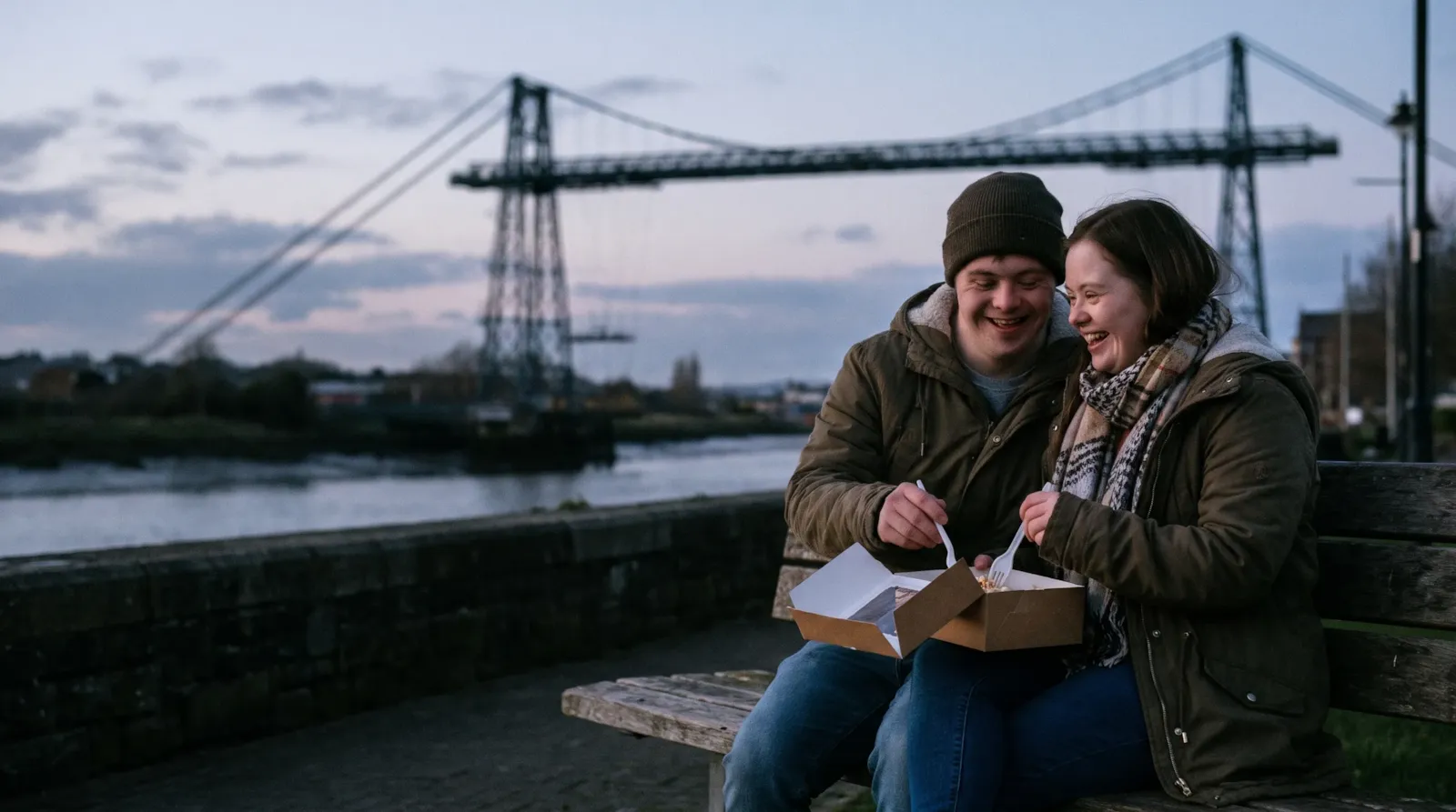 A young couple who both have Down syndrome sharing a cake on a riverside bench by the Newport Transporter Bridge