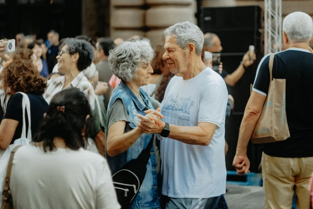 An older couple dancing together at a lively outdoor gathering while people around them watch and chat