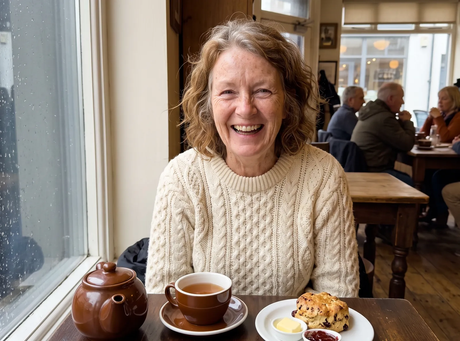A woman in her sixties with wavy auburn hair and a cream Aran jumper laughs at a cafe table by a rainy window, a pot of tea and a scone in front of her (AI-generated image)