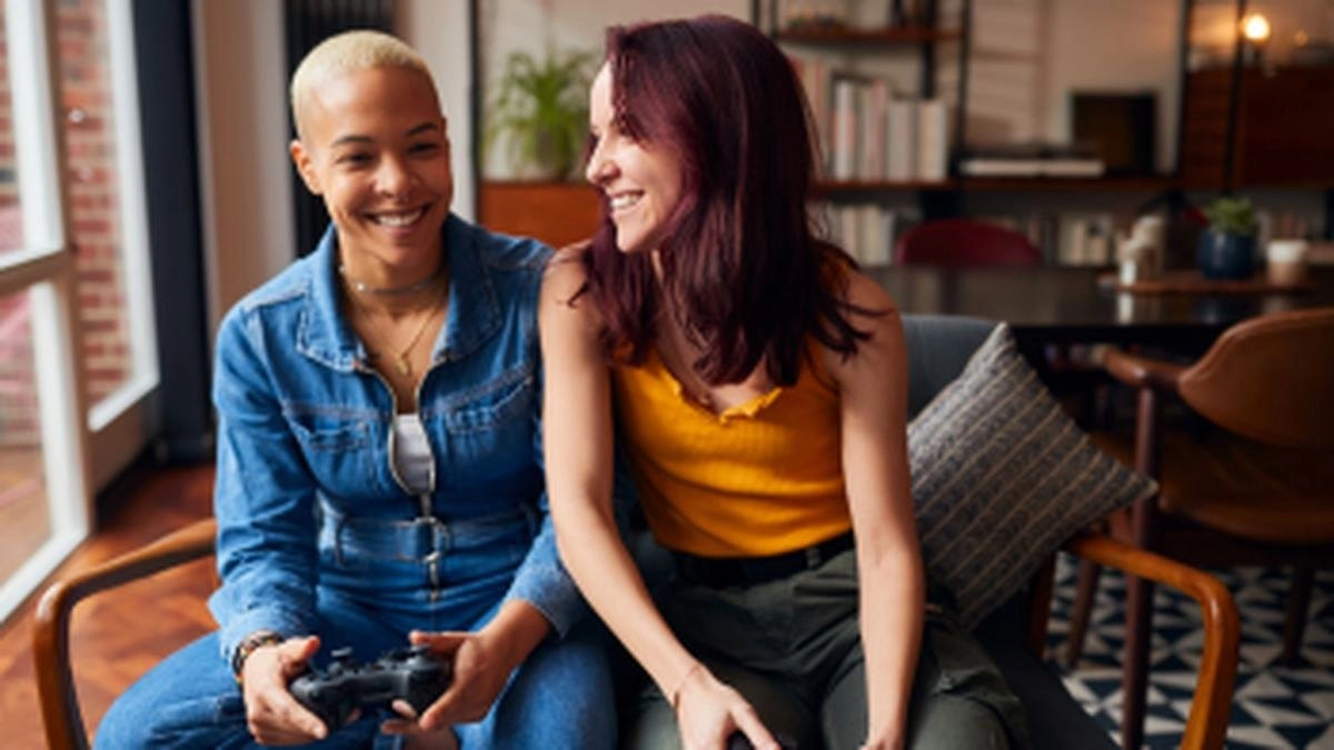 Two women laugh together on a sofa, one holding a game controller