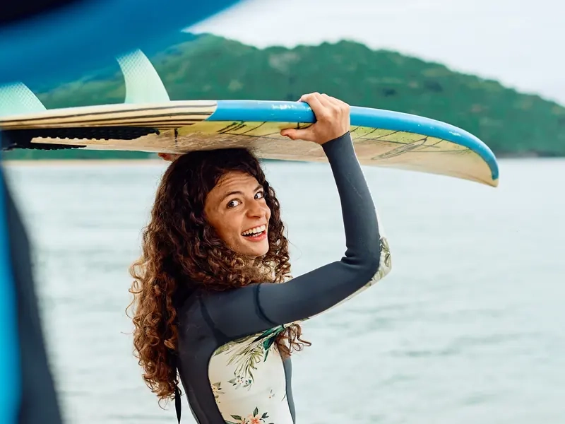 A smiling woman in a wetsuit carrying a surfboard on her head beside the water