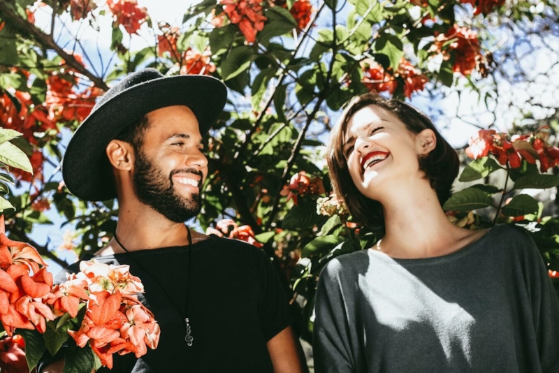 Una pareja se ríe bajo un árbol de flores rojas; él lleva sombrero y ella mira hacia arriba