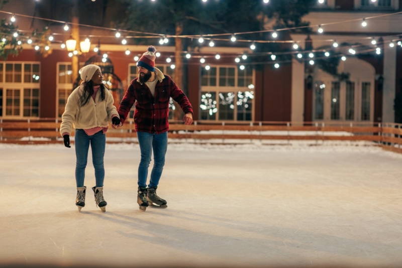 A couple holding hands and laughing on an outdoor ice rink under string lights