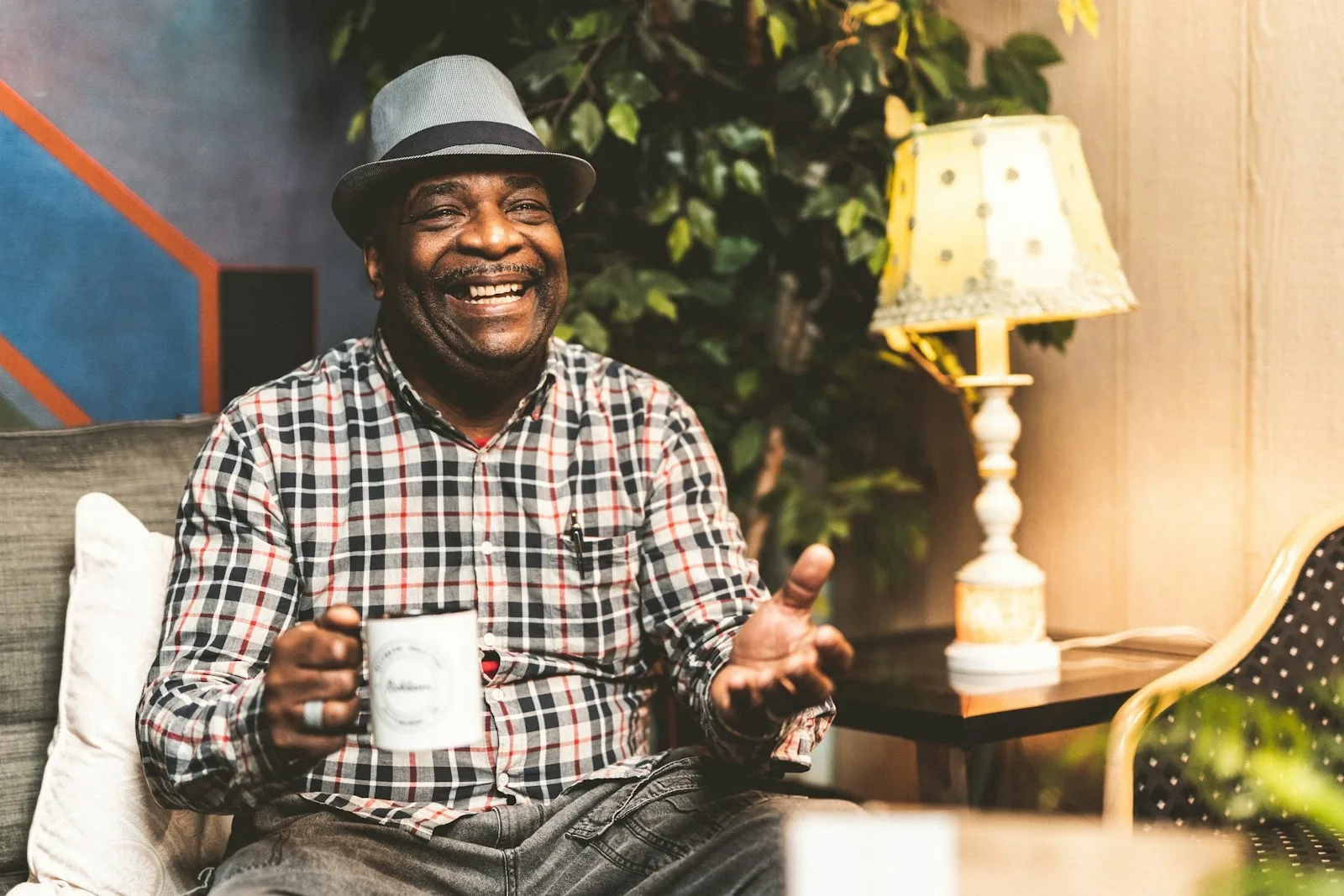 A smiling older man in a checked shirt and grey hat, mug in hand, chatting in a cosy room with a lamp glowing behind him