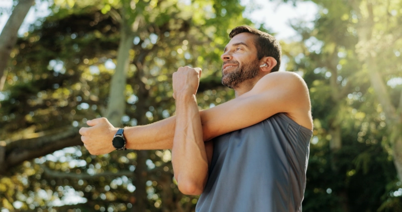 A smiling man stretching his arm before a run among sunlit trees