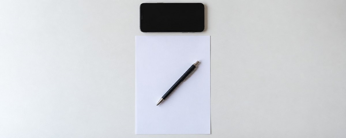 Seen from directly above, a blank sheet of paper and a pen on a white desk with a phone placed squarely along the top edge