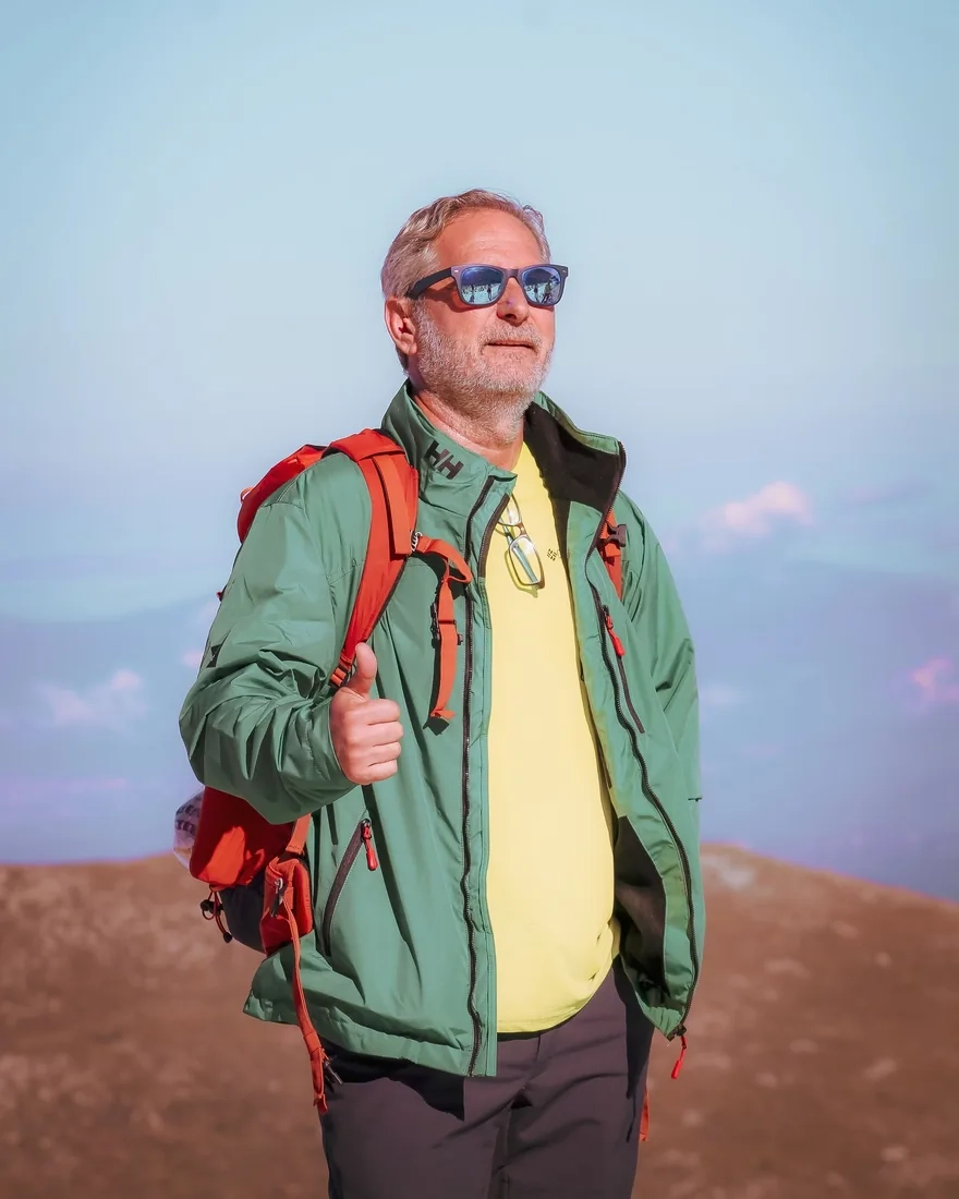 A man in sunglasses and a green jacket with a red backpack, standing on a sunny mountain ridge