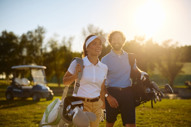 A smiling couple carrying golf bags across a sunlit course