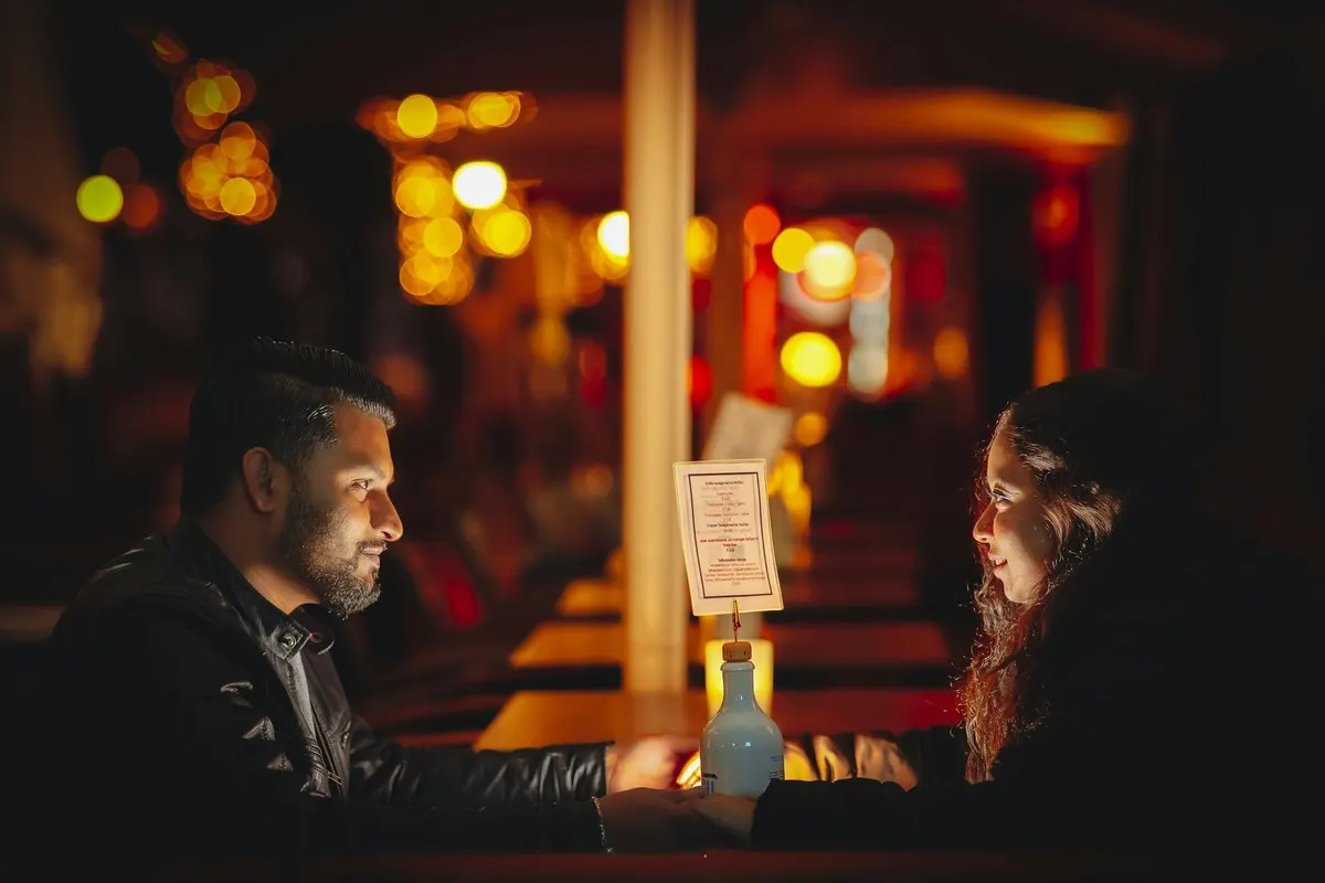 A bearded man and a woman with long curly hair talking across a candlelit table in a bar at night, warm lights blurred behind them