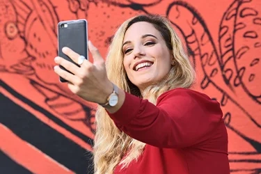 A young woman in a red top smiling as she takes a selfie in front of a painted mural