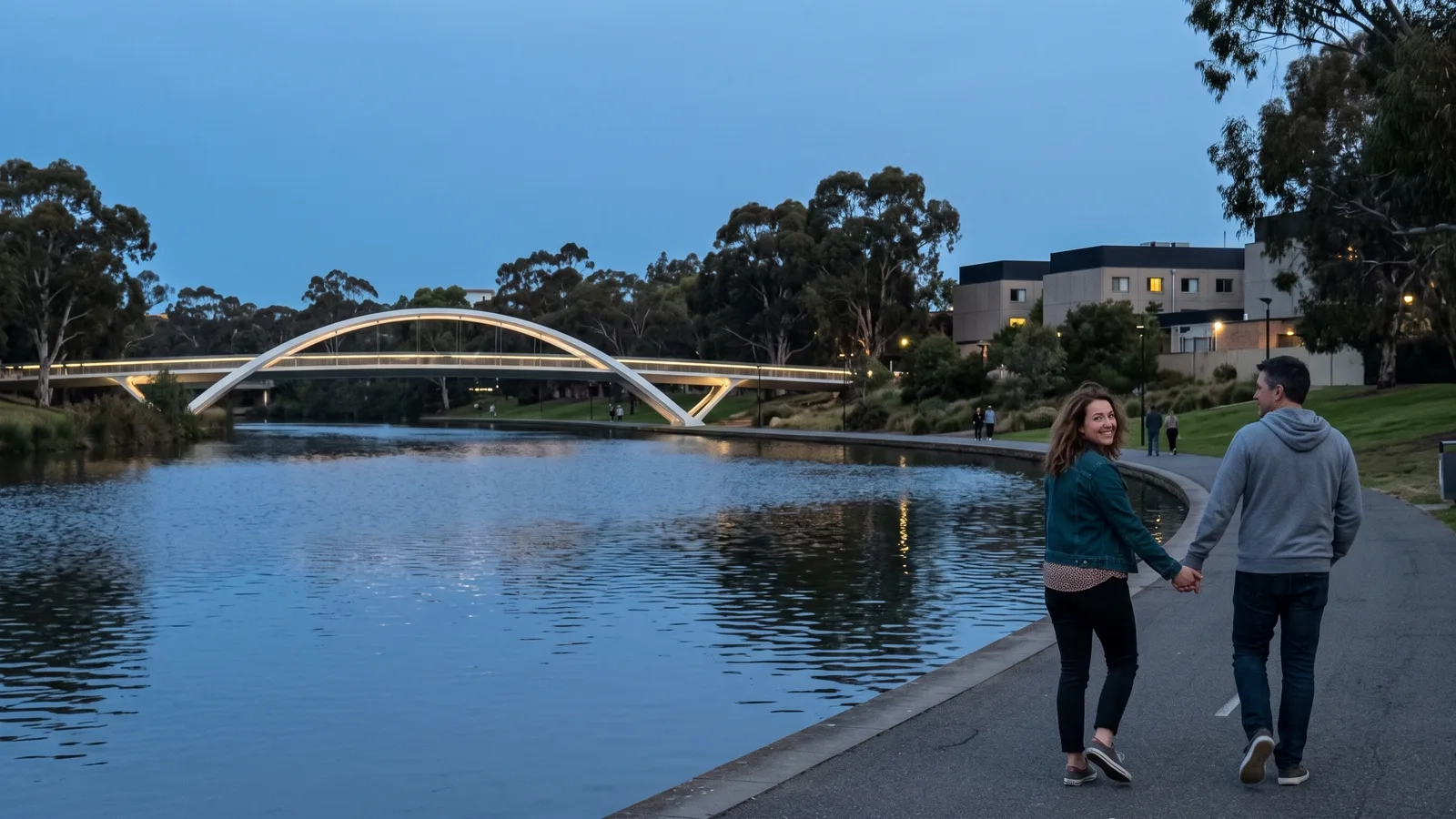 A couple walks hand in hand along the River Torrens in Adelaide at dusk, a lit footbridge and the city skyline behind them (AI-generated image)
