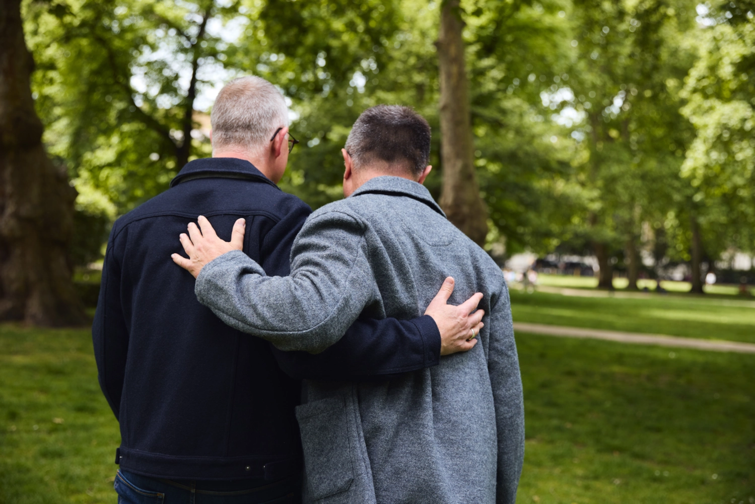Dos hombres mayores paseando por un parque, uno con el brazo sobre el hombro del otro.